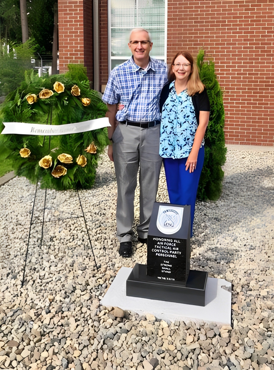 Couple standing beside a military memorial plaque and wreath.