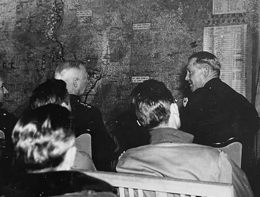 Military briefing with officers seated in front of a large tactical map.